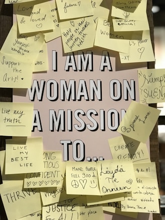A central poster with the text 'I AM A WOMAN ON A MISSION TO...' is surrounded by numerous sticky notes. Each note contains handwritten motivational messages or goals such as 'Support the Arts!', 'Love and be loved', 'Create beauty!', and 'Make people feel good'. The sticky notes are of various colors, mostly shades of yellow, and are adhered in a seemingly random pattern around the central poster.