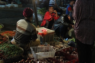 A bustling outdoor market scene at night featuring several people interacting and engaging with various goods. Vibrant baskets of red and green chilies are prominently displayed, surrounded by crates and boxes of other produce. The ambient light casts a warm glow over the activity, while some individuals are seated or crouched, examining or organizing items.