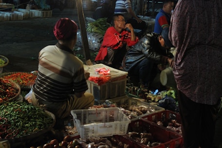 A bustling outdoor market scene at night featuring several people interacting and engaging with various goods. Vibrant baskets of red and green chilies are prominently displayed, surrounded by crates and boxes of other produce. The ambient light casts a warm glow over the activity, while some individuals are seated or crouched, examining or organizing items.