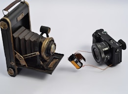A vintage accordion-style camera is placed on the left, facing a modern digital camera on the right. Between them lies a roll of photographic film, unspooled slightly, bridging the gap between the two devices. The setup is on a plain white background, highlighting the contrast between the old and new photographic technologies.