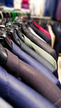 A row of neatly hung blazers on black plastic hangers. The blazers are in various shades, including blue, green, grey, and black. The focus is on the texture and material of the fabrics, with a vibrant, blurred background indicating a store setting.