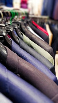 A row of neatly hung blazers on black plastic hangers. The blazers are in various shades, including blue, green, grey, and black. The focus is on the texture and material of the fabrics, with a vibrant, blurred background indicating a store setting.