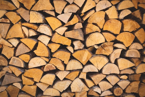 A neatly stacked pile of cut firewood pieces arranged in an organized manner, showcasing the cut wood grains and varying shades of brown and tan. The logs are cut into uniform lengths and are tightly packed together, creating a textured and patterned surface.