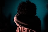 A person is obscured by a shadowy, dimly lit environment. Their head is lowered and covered by an arm, creating a sense of mystery and introspection against a teal background.