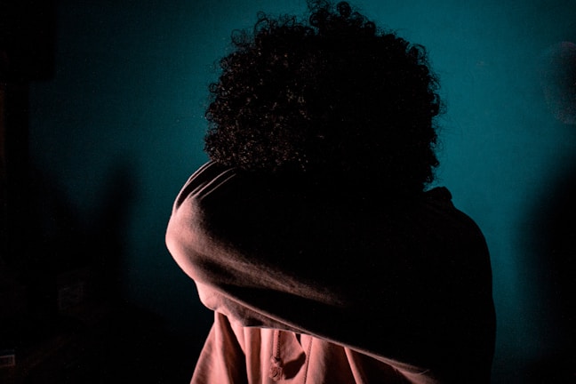 A person is obscured by a shadowy, dimly lit environment. Their head is lowered and covered by an arm, creating a sense of mystery and introspection against a teal background.