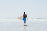 A surfer wearing bright Lapartisil board shorts, walking on a sunny beach with waves crashing behind.