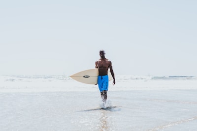 A surfer wearing bright Lapartisil board shorts, walking on a sunny beach with waves crashing behind.