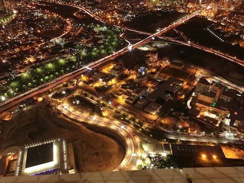A vibrant cityscape at night with illuminated streets and buildings. Roads form intricate patterns, with bright lights and heavy traffic. The city is surrounded by clusters of buildings, greenery, and visible infrastructure such as bridges and highways.
