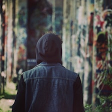 A close-up of a person wearing a dark denim jacket and a cozy hoodie, standing against a graffiti-covered city wall.