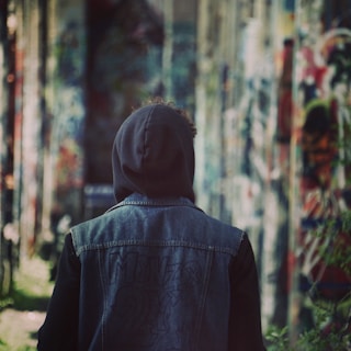 Urban scene featuring a young person wearing a dark hoodie and denim jacket, leaning against a graffiti wall.