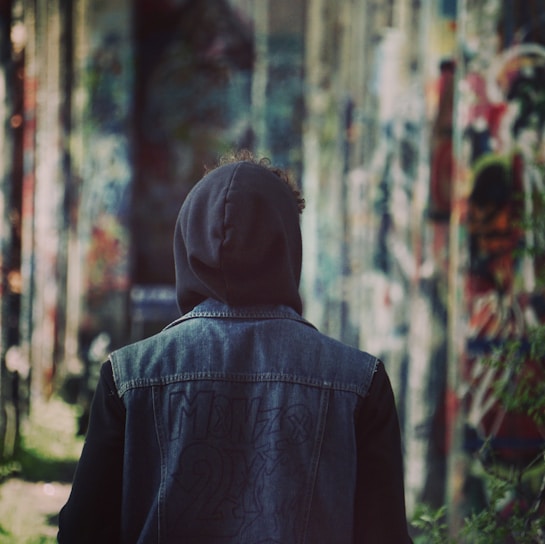 Urban scene featuring a young person wearing a dark hoodie and denim jacket, leaning against a graffiti wall.