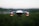 A small quadcopter drone is seen hovering in the air, equipped with a front-facing camera and red indicator lights. The background is slightly blurred, hinting at a natural landscape during a calm evening.
