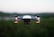A small quadcopter drone is seen hovering in the air, equipped with a front-facing camera and red indicator lights. The background is slightly blurred, hinting at a natural landscape during a calm evening.