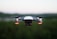 A small quadcopter drone is seen hovering in the air, equipped with a front-facing camera and red indicator lights. The background is slightly blurred, hinting at a natural landscape during a calm evening.