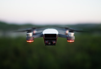 A small quadcopter drone is seen hovering in the air, equipped with a front-facing camera and red indicator lights. The background is slightly blurred, hinting at a natural landscape during a calm evening.