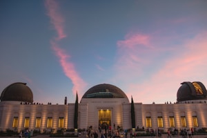 A large observatory building with a central dome flanked by two smaller domes is set against a twilight sky painted with soft pink and blue hues. People are seen gathered around and on the roof of the building, which is illuminated by warm lights from its windows.