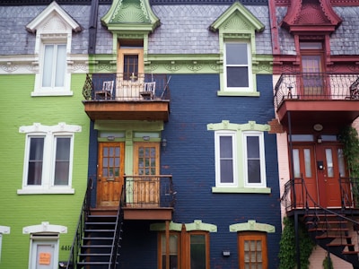 Three colorful urban row houses with distinct architectural features. The left house is painted in light green with white accents and a roof made of slate shingles. The middle building has a navy blue facade with green trims, featuring wooden doors and a small balcony with wrought iron railings. On the right, the house is maroon with more ornate designs and another iron balcony, surrounded by some climbing vines.