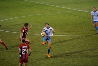 A soccer match with a player in a white shirt and blue shorts attempting to control the ball in mid-air. Opposing players in red and black uniforms are nearby, actively engaged in the play. The grass field is marked with lines, and teammates are positioned in the background.