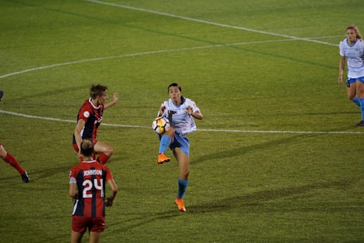 A soccer match with a player in a white shirt and blue shorts attempting to control the ball in mid-air. Opposing players in red and black uniforms are nearby, actively engaged in the play. The grass field is marked with lines, and teammates are positioned in the background.