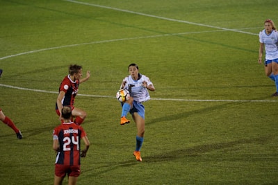 A soccer match with a player in a white shirt and blue shorts attempting to control the ball in mid-air. Opposing players in red and black uniforms are nearby, actively engaged in the play. The grass field is marked with lines, and teammates are positioned in the background.