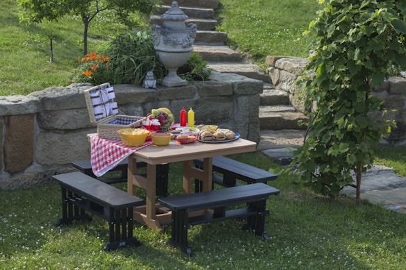 A picnic table set in a garden features a checkered cloth with a variety of foods including bowls of fruits, a basket with utensils, and condiments. The table is surrounded by benches on a grassy area, with stone steps and a decorative stone planter in the background. Lush greenery and flowers are visible around the picnic area, contributing to a serene outdoor setting.