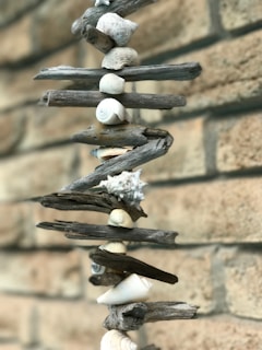 A vertical arrangement of driftwood pieces and seashells is displayed against a light brick wall. The driftwood and shells are alternated in a spiral pattern, creating a natural and artistic look.
