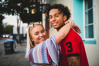 smiling woman with both hands wrap around man's neck near white concrete building