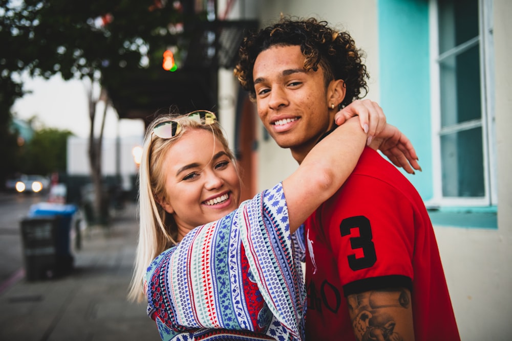 smiling woman with both hands wrap around man's neck near white concrete building