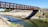 A wooden footbridge spans a narrow river or canal, supported by a concrete foundation on one side. The surrounding landscape includes dry, rocky banks with patches of green grass and shrubs. Trees are visible in the background under a clear blue sky.