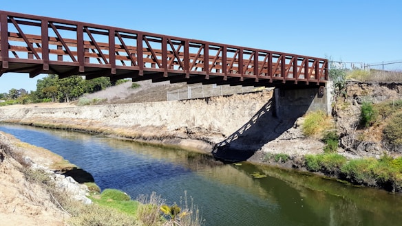 A wooden footbridge spans a narrow river or canal, supported by a concrete foundation on one side. The surrounding landscape includes dry, rocky banks with patches of green grass and shrubs. Trees are visible in the background under a clear blue sky.