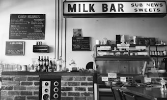A black and white photo of a vintage-style milk bar with a brick counter. On the counter, there are various bottles and a speaker. A menu board lists cold drinks, milkshakes, and juices. Above, a sign reads 'MILK BAR' along with other words like 'SUB NEWS' and 'SWEETS'. The background shows shelves lined with products and a display case containing food items.