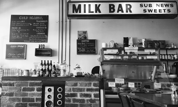 A black and white photo of a vintage-style milk bar with a brick counter. On the counter, there are various bottles and a speaker. A menu board lists cold drinks, milkshakes, and juices. Above, a sign reads 'MILK BAR' along with other words like 'SUB NEWS' and 'SWEETS'. The background shows shelves lined with products and a display case containing food items.