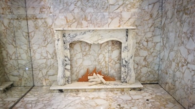 An ornate marble fireplace set against a heavily veined marble wall. The fireplace features intricate carvings and columns. On the hearth, there are log-shaped decor items with a stylized orange flame designed as part of the display.