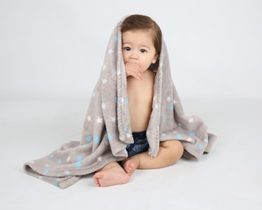 A young child sits wrapped in a soft, gray blanket adorned with blue and white stars. The child has a thoughtful expression and seems to be sucking on their thumb. The background is a plain, light color, enhancing the cozy and gentle atmosphere.
