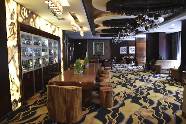 A luxurious interior space with a long wooden table flanked by unique wooden stools. The area features intricate lighting fixtures, including chandeliers, and showcases a patterned carpet with organic designs. A display cabinet filled with various objects lines one wall, while comfortable seating areas are arranged throughout the space.
