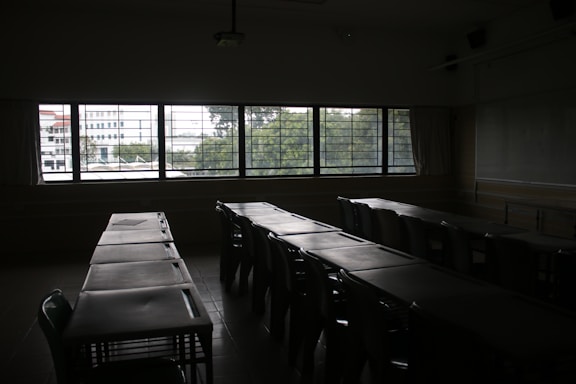 A dimly lit classroom with rows of empty desks and chairs facing a large window. Through the window, an outside view of trees and buildings is partially visible. The room appears quiet and unoccupied.