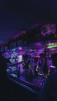 A vibrant neon-lit bar interior with colorful lights reflecting off the polished wooden counter, lively patrons enjoying drinks and laughter filling the air.