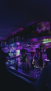 Dimly lit bar interior with vibrant neon lights. Bartenders work behind a shiny counter filled with bottles and glasses. The ambient light casts a purple and blue hue throughout the space, creating a lively and atmospheric setting.
