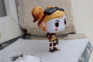 A small, stylized figurine with a large head and exaggerated features stands on a snowy surface. The figure has orange hair tied in pigtails, wears goggles on its head, and is dressed in a brown and red outfit with boots. It carries a small object resembling a gun. The background is a textured wall, and the area around the figurine is covered in a light layer of snow.