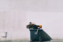 A large green dumpster is positioned against a light-colored concrete wall. Overflowing with various pieces of cardboard and debris, the dumpster casts a noticeable shadow across the ground.