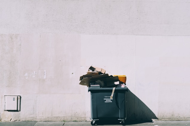 A large green dumpster is positioned against a light-colored concrete wall. Overflowing with various pieces of cardboard and debris, the dumpster casts a noticeable shadow across the ground.