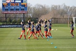 A group of female soccer players in blue jerseys and red shorts are jogging on a grassy field. Training cones are placed on the ground. In the background, a large digital scoreboard shows the time and score. Bare trees and a fence line the field's edge.