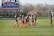 A group of female soccer players in blue jerseys and red shorts are jogging on a grassy field. Training cones are placed on the ground. In the background, a large digital scoreboard shows the time and score. Bare trees and a fence line the field's edge.