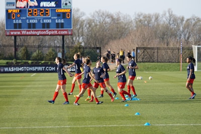 A group of female soccer players in blue jerseys and red shorts are jogging on a grassy field. Training cones are placed on the ground. In the background, a large digital scoreboard shows the time and score. Bare trees and a fence line the field's edge.