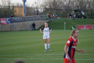 A soccer player in a white uniform stands on a grass field with hands on hips, looking forward. Another player in a red and blue uniform appears in the foreground, slightly out of focus. There is a fence and spectators on a hill in the background, along with a vehicle and banners.