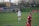 A soccer player in a white uniform stands on a grass field with hands on hips, looking forward. Another player in a red and blue uniform appears in the foreground, slightly out of focus. There is a fence and spectators on a hill in the background, along with a vehicle and banners.