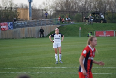 A soccer player in a white uniform stands on a grass field with hands on hips, looking forward. Another player in a red and blue uniform appears in the foreground, slightly out of focus. There is a fence and spectators on a hill in the background, along with a vehicle and banners.