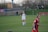 A soccer player in a white uniform stands on a grass field with hands on hips, looking forward. Another player in a red and blue uniform appears in the foreground, slightly out of focus. There is a fence and spectators on a hill in the background, along with a vehicle and banners.