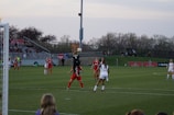 A soccer match is taking place on a grass field with players in red and black, and white uniforms. A player in a black jersey is jumping to catch a ball near the goalpost, while other players are observing or preparing to move. The stands in the background are filled with spectators, and trees line the horizon under a slightly overcast sky.