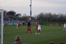A soccer match is taking place on a grass field with players in red and black, and white uniforms. A player in a black jersey is jumping to catch a ball near the goalpost, while other players are observing or preparing to move. The stands in the background are filled with spectators, and trees line the horizon under a slightly overcast sky.